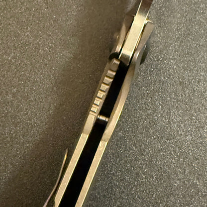 Close-up of Strider SnG CC compound grind knife showing blade spine, full Acid Bronze titanium scales, and open-back construction.