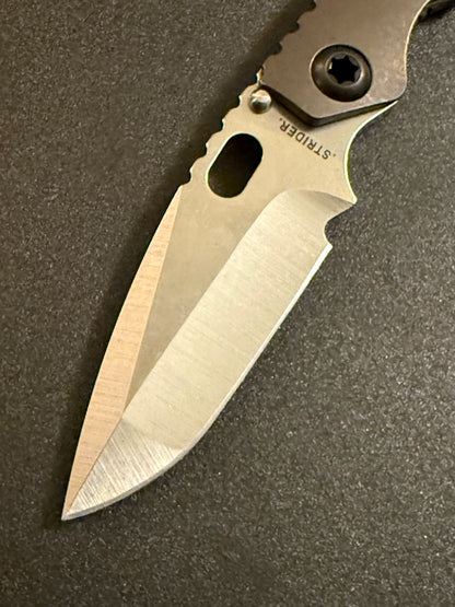 Detailed Strider SnG CC CPM MagnaCut compound grind blade with contrasting satin bevels, oval thumb opening, and Acid Bronze titanium handle.