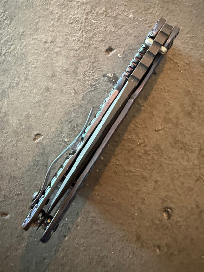Rear spine view of Strider SMF Monkey Edge FRAG showing open titanium frame, pocket clip and textured heat-anodized handle scales.