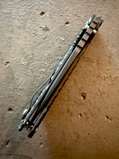 Rear spine view of Strider SMF full titanium DGG folding knife displaying frame construction, pocket clip, blade centering and Double Gunner Grip handle texture.