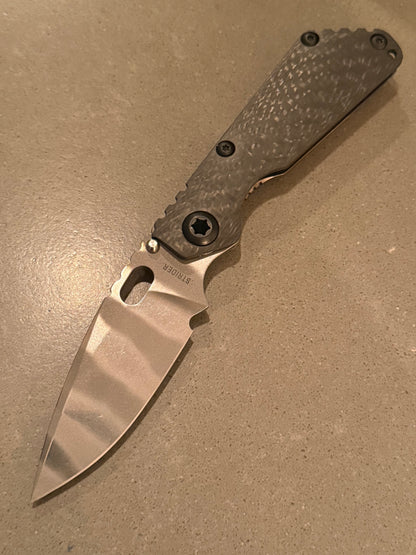 Strider SnG CC MagnaCut folding knife with Ghost Stripe blade finish, textured carbon fiber handle, and upgraded titanium hardware.